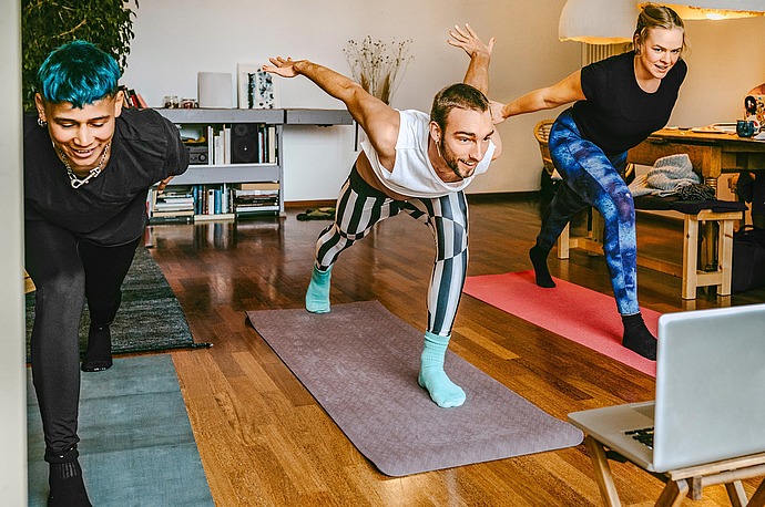 Drei Personen üben drinnen gemeinsam Yoga. Sie balancieren auf einem Bein mit ausgestreckten Armen und folgen den Anweisungen eines Laptops auf dem Boden. Sie lächeln und scheinen die Sitzung zu genießen.