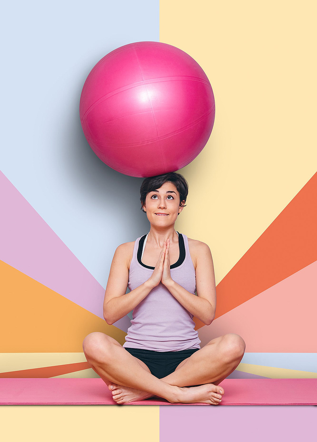 Eine Frau sitzt im Schneidersitz auf einer Yogamatte, hält die Hände in Gebetshaltung und balanciert einen großen rosafarbenen Gymnastikball auf ihrem Kopf. Im Hintergrund sind bunte geometrische Formen zu sehen.