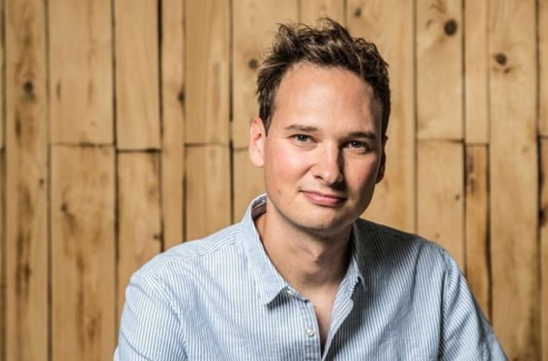 Ein Mann mit kurzen braunen Haaren, einem hellblau gestreiften Hemd und Jeans sitzt mit gefalteten Händen auf einem Stuhl vor einer Holzpaneelwand und lächelt leicht in die Kamera.